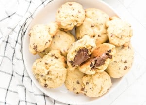 A plate of chocolate chip cookies, with one cookie broken in half to reveal a melted chocolate filling, placed on a white surface next to a checkered cloth.