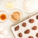 Baking ingredients in bowls, including eggs, butter, brown sugar, flour, chocolate chips, and vanilla, alongside a tray of chocolate-covered treats on parchment paper.