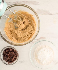 Overhead view of a mixing bowl with cookie dough and a hand mixer, next to bowls of chocolate chips and flour on a white surface.