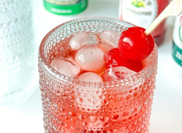 A textured glass filled with ice and red liquid, garnished with two maraschino cherries on a cocktail pick.