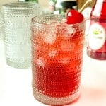 A textured glass filled with a red iced drink, garnished with a maraschino cherry on top; bottles and another glass are in the background.