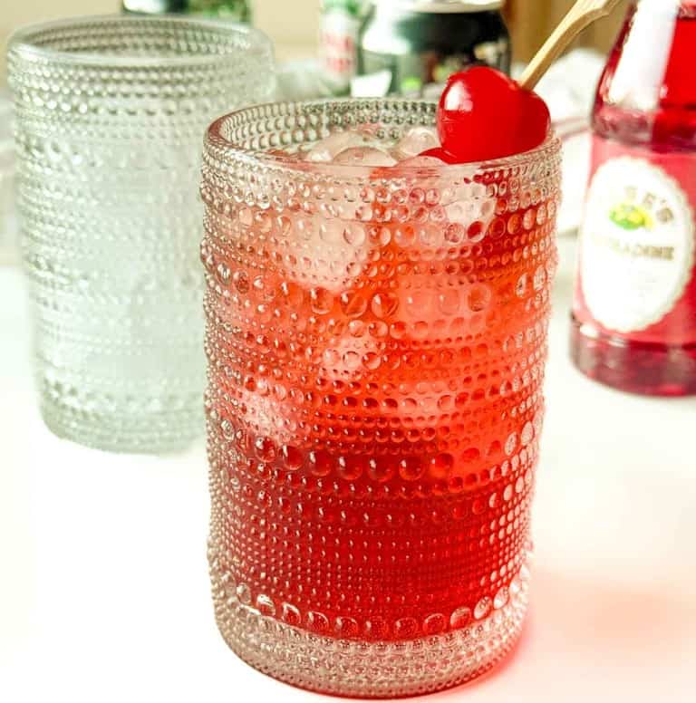 A textured glass filled with a red iced drink, garnished with a maraschino cherry on top; bottles and another glass are in the background.