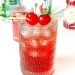 A textured glass filled with ice and a red drink, garnished with two maraschino cherries on a skewer; 7UP and grenadine bottles are in the background.