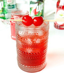 A textured glass filled with ice and a red drink, garnished with two maraschino cherries on a skewer; 7UP and grenadine bottles are in the background.
