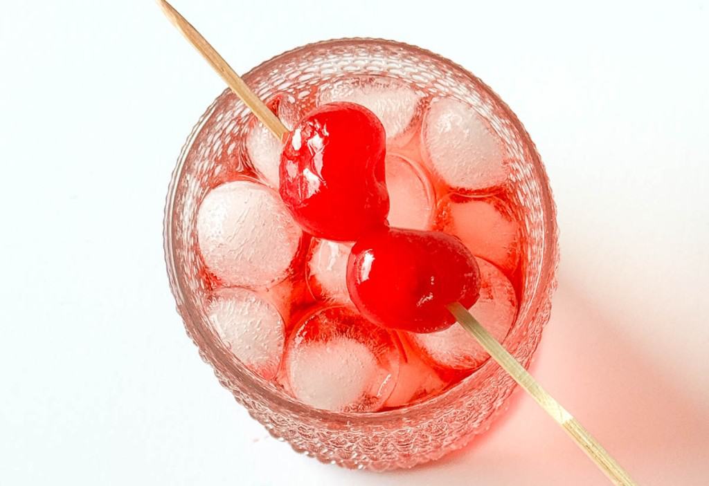 A glass filled with ice cubes and a red beverage, garnished with two maraschino cherries on a wooden skewer.