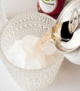 A can of soda is being poured over ice cubes in a textured glass, with a bottle of grenadine visible in the background.