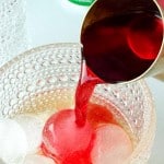 A metal cup pours red liquid over ice cubes in a textured glass, with a sugar jar visible in the background.