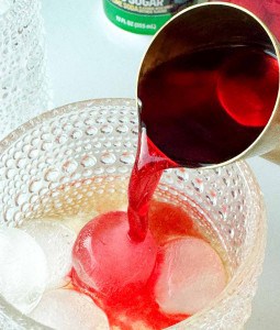 A metal cup pours red liquid over ice cubes in a textured glass, with a sugar jar visible in the background.