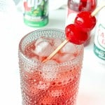 A textured glass filled with ice and a red drink, garnished with two maraschino cherries on a skewer. Bottles and cans of soda are visible in the background.