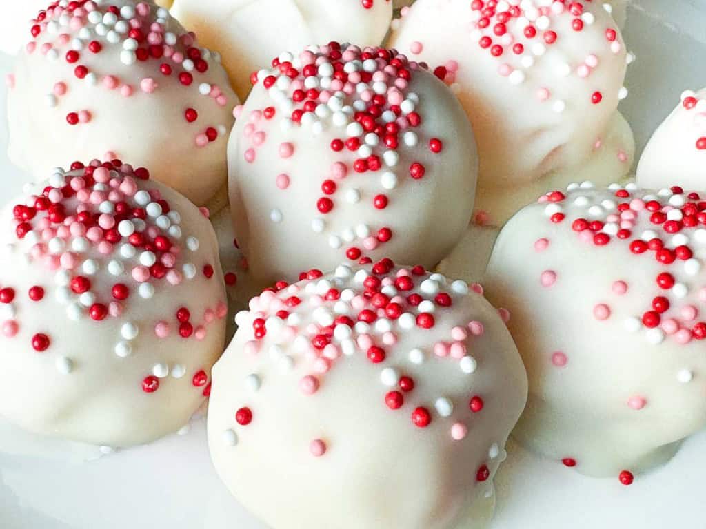 Close-up of white chocolate-coated cake balls topped with red, pink, and white sprinkles, arranged on a white surface&mdash;perfect Valentine's Day treats for someone special.