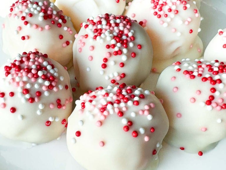 Close-up of white chocolate-coated cake balls topped with red, pink, and white sprinkles, arranged on a white surface—perfect Valentine's Day treats for someone special.