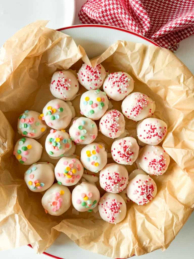 A plate lined with parchment paper holds round white chocolate-coated cake balls topped with colorful sprinkles and red sugar crystals—perfect Valentine's Day treats.