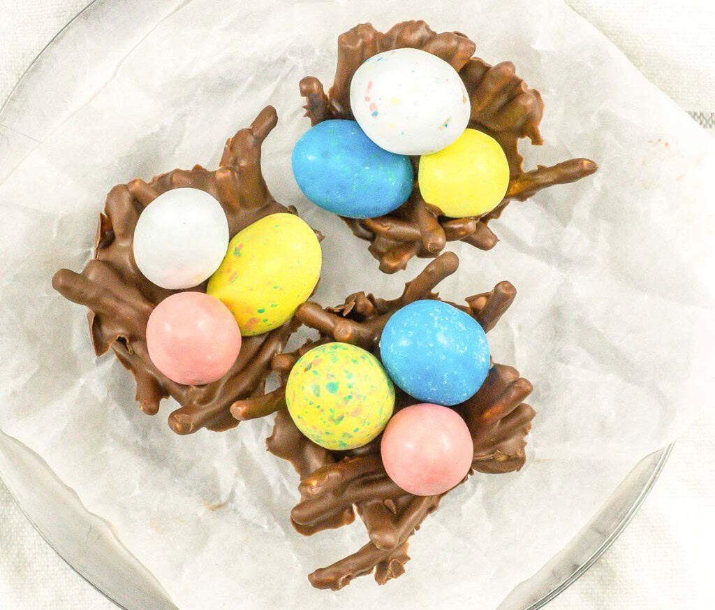 Three chocolate nests topped with colorful candy eggs are arranged on parchment paper.