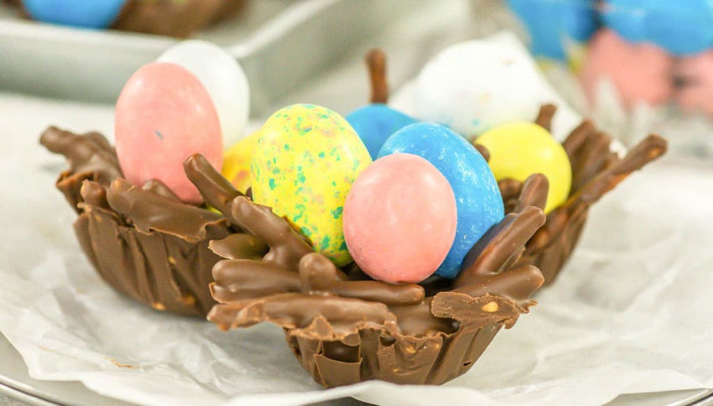 A chocolate nest made of chocolate-covered pretzel sticks holds colorful candy eggs on a piece of parchment paper.