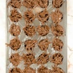 A metal muffin tin lined with plastic wrap contains clusters of chocolate-covered stick-shaped snacks, arranged in each section.