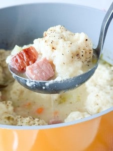 A ladle lifting ham, vegetables, and a dumpling from a pot of creamy soup.