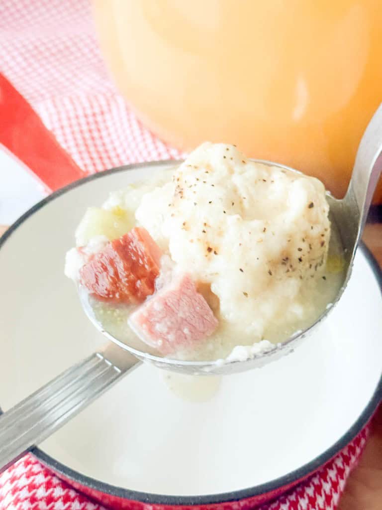 A ladle of creamy soup with dumplings, chunks of sausage, and vegetables held over a white bowl on a red and white cloth.