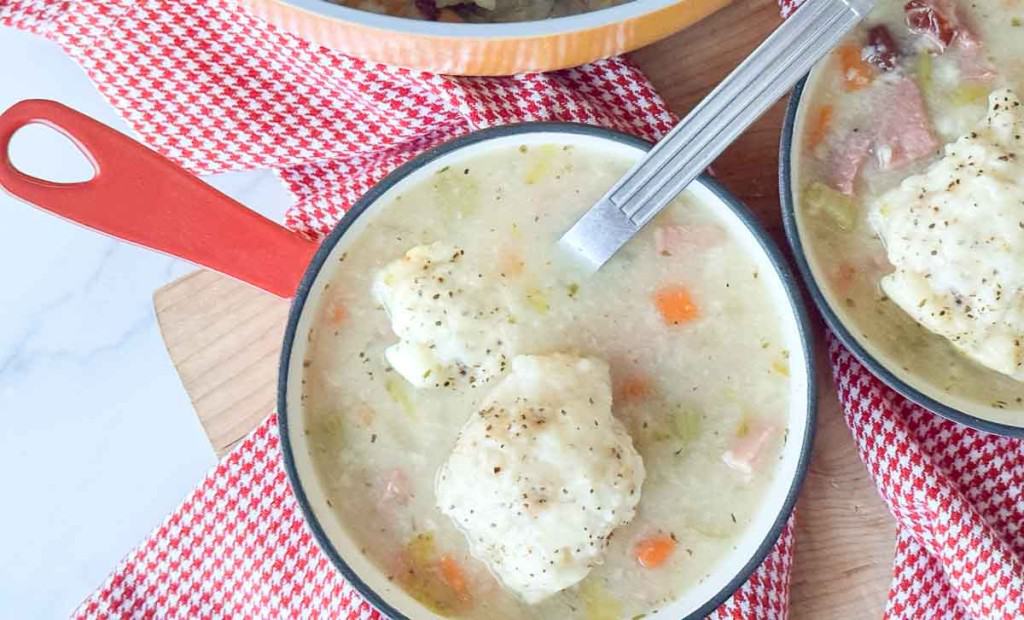 A bowl of creamy soup with vegetables, ham, and a dumpling, served with a spoon on a red and white checkered cloth.