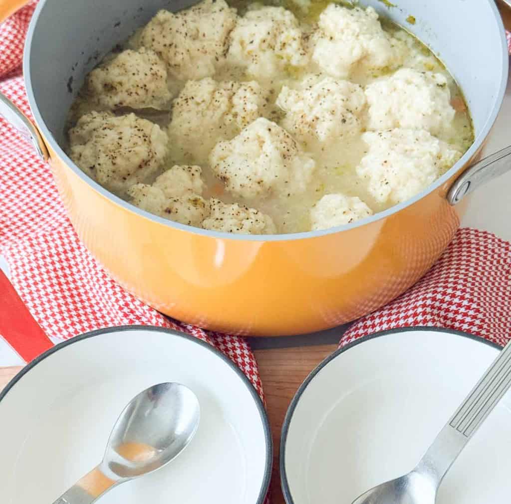 A yellow pot filled with broth and dumplings sits on a red and white cloth, with two empty bowls and spoons beside it.