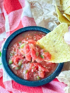 A tortilla chip is dipped into a bowl of chunky tomato salsa, with a red and white checkered cloth in the background.
