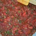 Chopped tomatoes, green peppers, and onions mixed in a pan with a yellow spatula and wooden handle visible at the top right.
