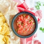A black bowl of chunky tomato salsa sits on a red and white cloth next to a pile of tortilla chips and sprigs of cilantro.