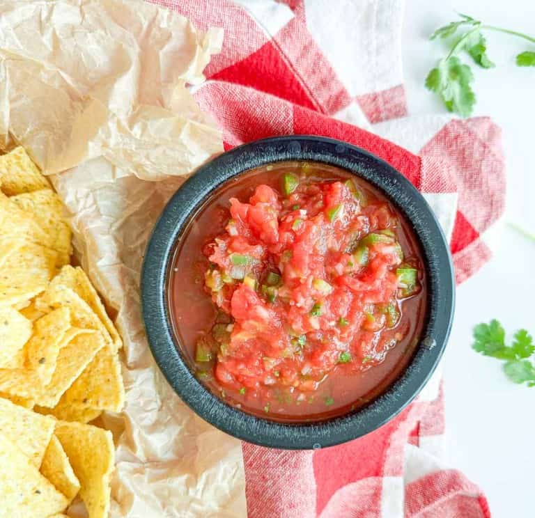 A black bowl of chunky tomato salsa sits on a red and white cloth next to a pile of tortilla chips and sprigs of cilantro.
