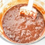 A glass mixing bowl filled with chocolate brownie batter is being stirred with a white spatula.