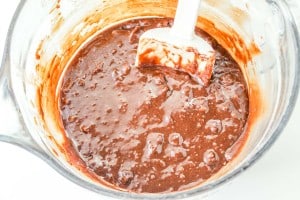 A glass mixing bowl filled with chocolate brownie batter is being stirred with a white spatula.
