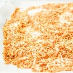 A baking sheet lined with parchment paper holds an even layer of toasted shredded coconut. A can with an orange label is partially visible in the background.