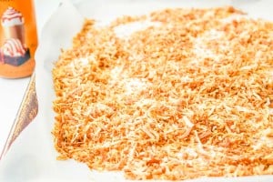 A baking sheet lined with parchment paper holds an even layer of toasted shredded coconut. A can with an orange label is partially visible in the background.