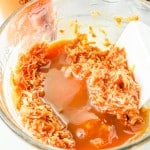 A glass bowl containing shredded coconut mixed with caramel sauce, being stirred with a white spatula.
