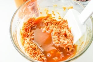A glass bowl containing shredded coconut mixed with caramel sauce, being stirred with a white spatula.