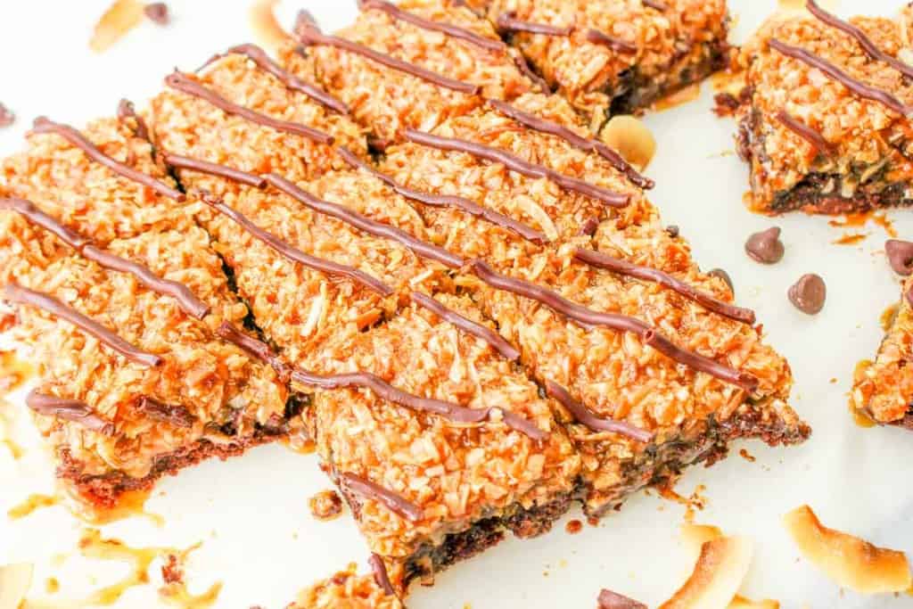 Rectangular dessert bars topped with toasted coconut and drizzled with chocolate, arranged on a white surface with scattered chocolate chips and coconut flakes.