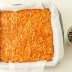 A square baking pan lined with parchment paper contains an unbaked oat mixture. A small bowl of chocolate chips is placed beside the pan.