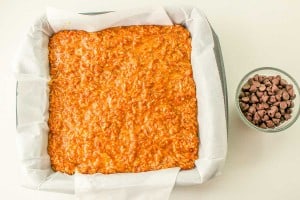 A square baking pan lined with parchment paper contains an unbaked oat mixture. A small bowl of chocolate chips is placed beside the pan.
