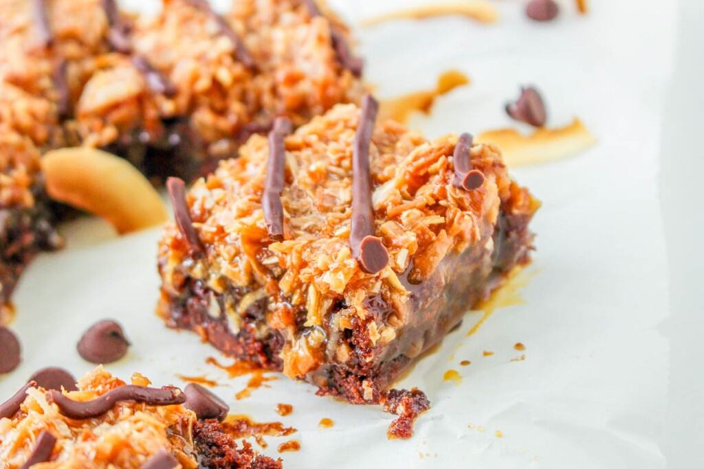A close-up of a dessert bar with a chocolate base, caramelized coconut topping, and chocolate drizzle, set on parchment paper with scattered chocolate chips.