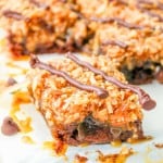 A close-up of caramel and coconut dessert bars topped with a drizzle of chocolate and chocolate chips, arranged on a white surface.