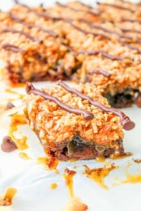 A close-up of caramel and coconut dessert bars topped with a drizzle of chocolate and chocolate chips, arranged on a white surface.