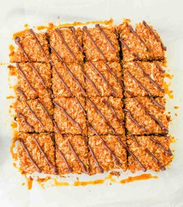 A batch of sixteen caramel coconut bars with chocolate drizzle, arranged in a grid on parchment paper.