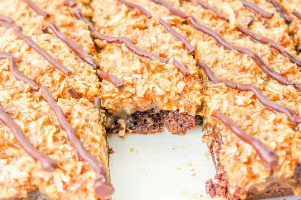 A close-up of caramel coconut brownies topped with toasted coconut and chocolate drizzle, with one piece missing.