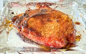 A baked ham with a golden-brown glaze sits on a sheet of aluminum foil.