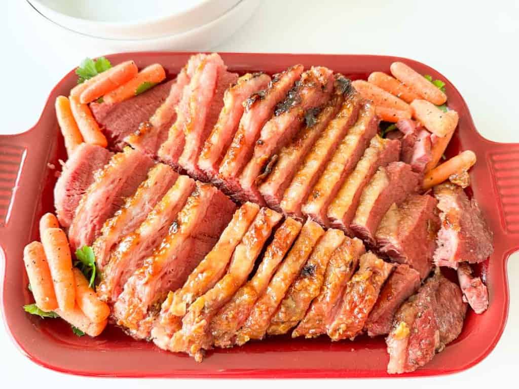 Sliced corned beef arranged on a red platter, garnished with baby carrots and fresh parsley.