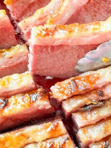 Close-up of sliced, glazed slow cooker corned beef being served with metal tongs. The meat is pink with a caramelized exterior.