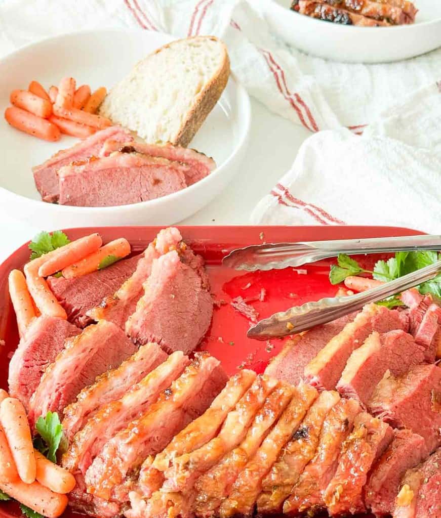 Sliced slow cooker corned beef and cooked carrots on a red tray with tongs, served with bread and more carrots on a white plate in the background.