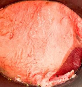 A large cut of raw corned beef with a thick layer of fat is shown in a cooking pot with visible liquid and spices.
