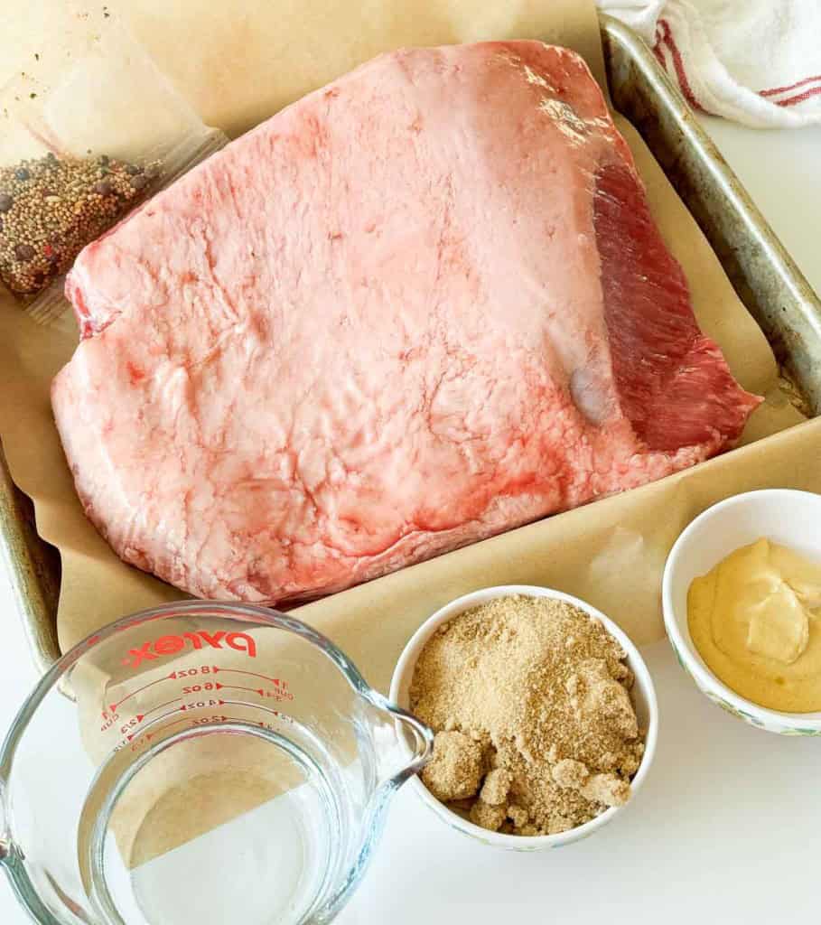 Raw brisket on a parchment-lined baking sheet, next to bowls of brown sugar, mustard, pickling spices, and a glass measuring cup with water&mdash;perfect ingredients for homemade slow cooker corned beef.