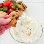 A hand dips a chocolate chip cookie into a bowl of creamy white dip, with strawberries, pretzels, and cookies in the background on a white surface.