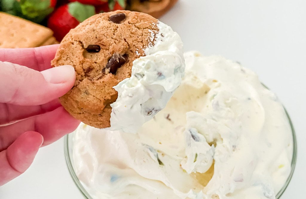 A hand holds a chocolate chip cookie dipped in a bowl of creamy white dessert.