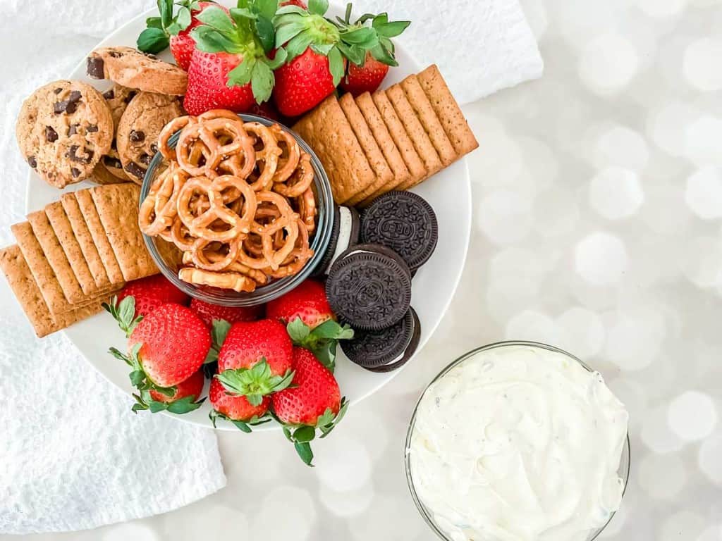A plate with strawberries, pretzels, assorted cookies, and crackers, next to a bowl of creamy dip on a light-colored surface.